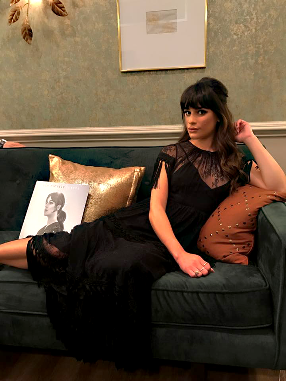 Image of Lea Michele sitting on a couch and looking at the camera. Besides her is her latest album on Vinyl.